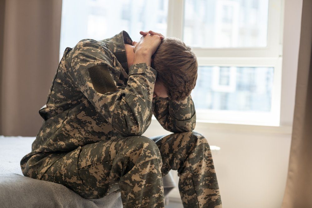 Person in a digital camouflage uniform sitting on the edge of a bed with head in hands, beside a bright window