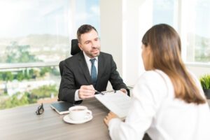 A lawyer and his client sit at his desk. Over a cup of coffee, he explains what the VA DBQ is and hands her the form.