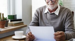 Older man with glasses and a gray beard reading a letter at a wooden table by a window