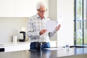 Older man in glasses reading documents while standing at a kitchen counter
