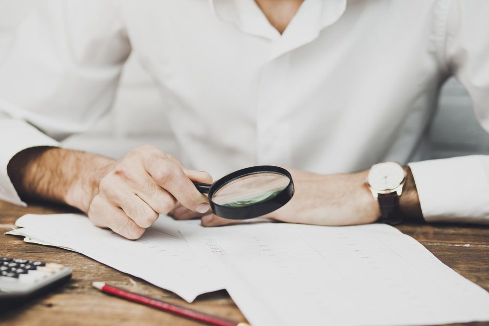 Man in a white shirt examining printed documents with a magnifying glass at a wooden desk, with a calculator and pencil nearby